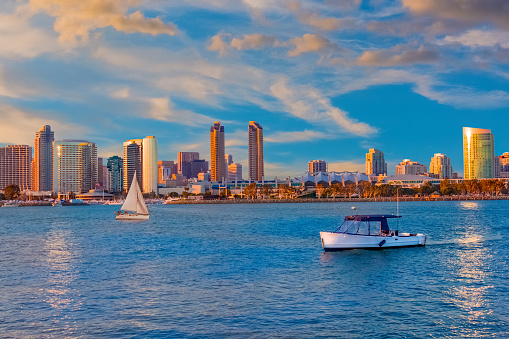San Diego Bay glows in golden dusk ,  and the light is reflected off of the skyline's buildings. Two boats sail in the harbor, in the evening twilight.
