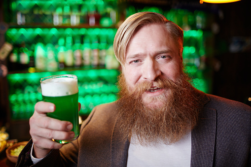 Cheerful bearded man toasting with glass of green foaming beer in pub