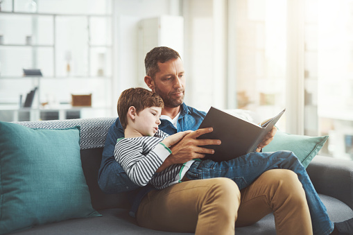 Shot of a carefree little boy and his father relaxing on a sofa while reading a book at home during the day