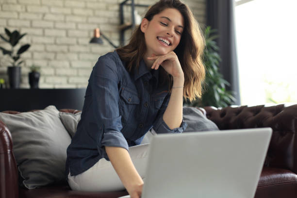 istock Smiling young woman sitting on sofa with laptop computer and chating with friends. 1253012630