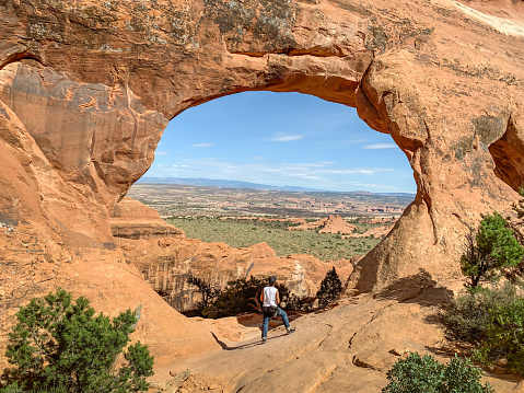Amazing view of distant rock mountains with a natural window through  the hole in a window on the walls of red clay rock. hiker young man is standing looking out of rock frame at Arches national park