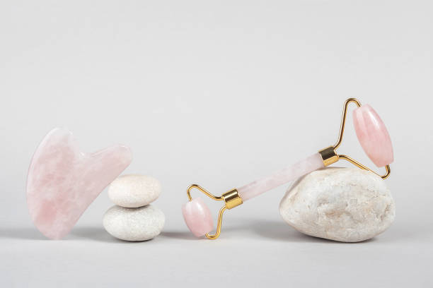 istock Crystal rose quartz facial roller and massage tool jade Gua sha on stones, grey background. Facial anti-age massage for natural lifting and toning treatment at home. Beauty concept Front view 1253013722