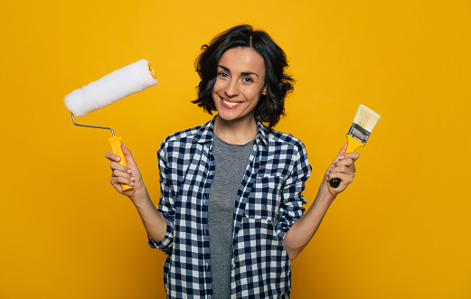Half-length photo of a smiling nice girl, in a black and white checkered shirt, holding a paint roller in her right hand and a brush in her left hand.