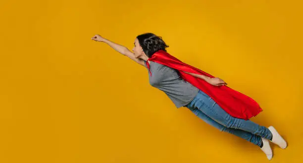 I am flying! A full-length, turned a left photo of a young girl, portraying a flying superheroine, that is in a classic superhero pose, dressed casually, but with a red superhero fluttering cloak.