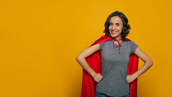 I am a superwoman! A young, beautiful girl, standing confidently with her fists on her waist, wearing red superman cloak, smiling like a real hero, ready to accomplish any task in the world.