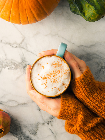 Autumn flat lay with mug of latte coffee on marble table with female hands in cozy orange sweater holding the cup. Cozy fall