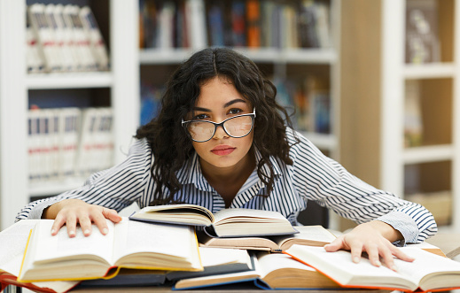 Exhaustion Concept. Frustrated hispanic female student in specs sitting at desk, leaning on books at library, copyspace