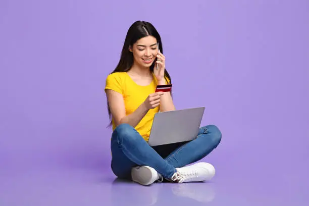 Photo of Asian girl talking on cellphone, making order with laptop and credit card