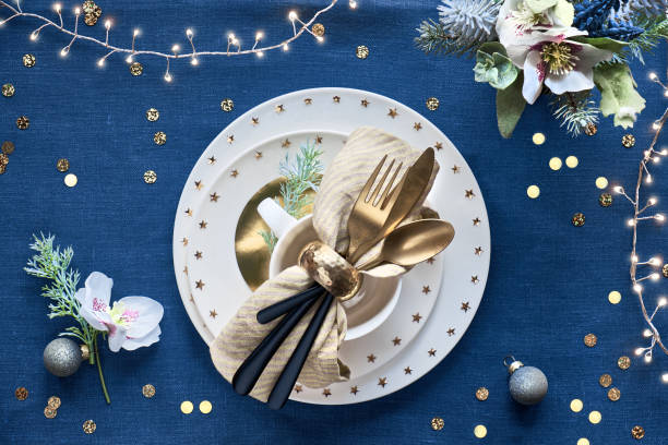 istock Christmas table setup with white plate and golden utensils and gilded decorations. Flat lay on classic blue linen textile background. Xmas lights garland. 1253024181