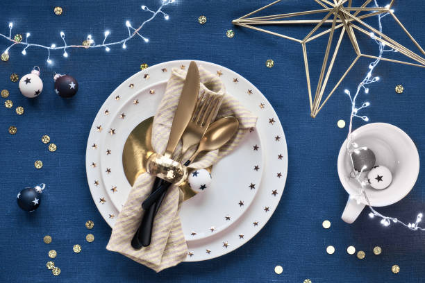 istock Christmas table setup with white plate and golden utensils and gilded decorations. Flat lay, top view on classic blue. Dark blue linen textile background. Xmas lights garland. 1253024284