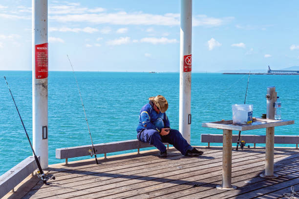 Townsville, Queensland, Australia - June 2020: A fisherman playing with his phone while he sits at the end of a jetty with his rods out in the water