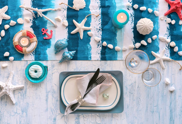 istock Columbus Day lunch table setting with nautical sea decorations on blue stripy runner, classic blue white plates, utensils on white rustic table. Top view, casual modern lunch table setup. 1253026711