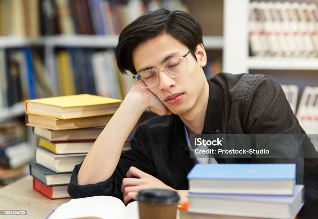 Tired student napping in the university library Exam Time. Exhaused japanese student in eyewear sleeping in the library among stacks of many books, copy space Asian and Indian Ethnicities Stock Photo