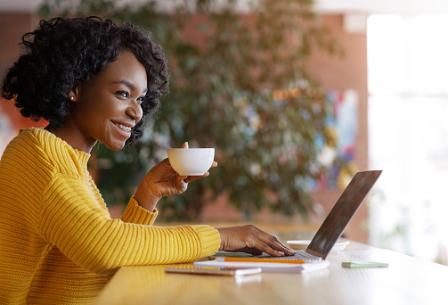 Pretty black woman freelancer drinking coffee and smiling to someone, working with laptop at cafe, free space