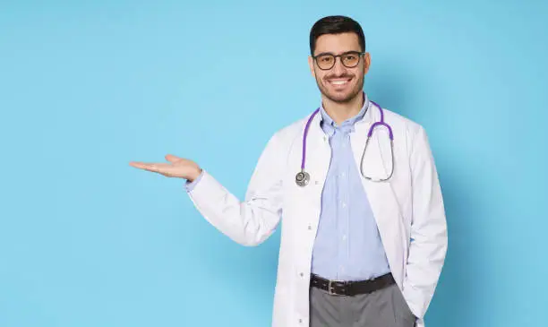 Photo of Horizontal banner of smiling young male doctor standing in medical robe and eyeglasses, showing commercial offer with hand, copy space on left, isolated on blue background