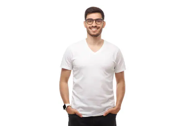 Photo of Young man standing with hands in pockets, wearing glasses and blank white tshirt with copy space, isolated on studio background