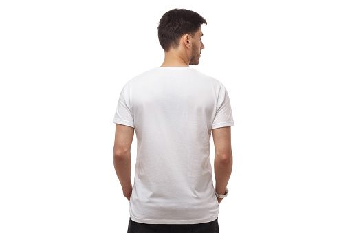 Back view of young man standing with hands in pockets, wearing blank t shirt with copy space, isolated on white studio background