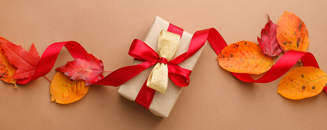 Autumn creative holiday present. Handmade paper gift box with satin bow, foliage dried leaves on beige background. Thanksgiving day, fall background. discount, promotion concept