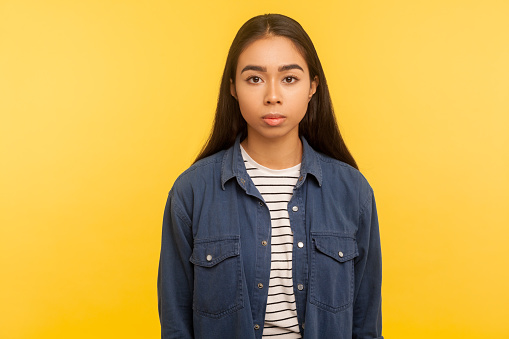 Portrait of serious unsmiling brunette girl in denim shirt standing calm, looking focused concentrated at camera, earnest attentive expression. indoor studio shot isolated on yellow background