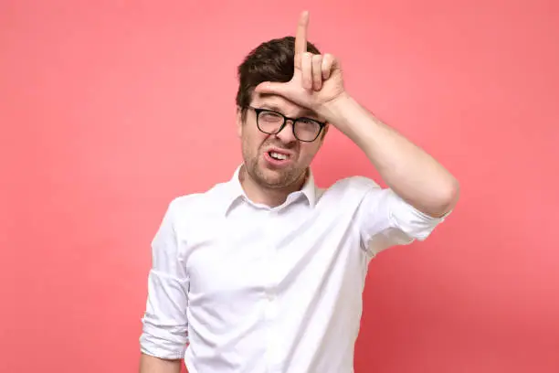 Young caucasian boy in glasses showing looser sign. Studio shot on red wall.