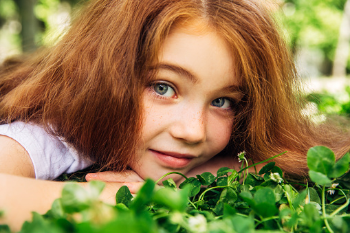 A red-haired little girl in a green T-shirt sits near the window at home and smiles. Portrait of a cute five year old girl
