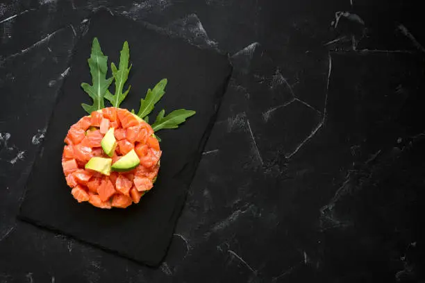 Raw salmon tartare. Fresh salmon, avocado and arugula salad in a culinary ring on a slate plate, black stone background. Top view, flat lay, copy space