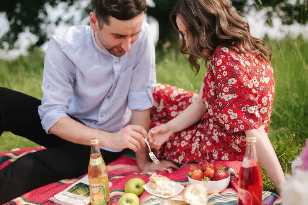 Happy couple spend time outdoors on mini picnic. Attractive woman and handsome man sits on red blanker by the big tree.