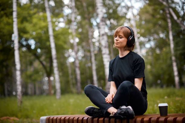 音楽を聴いて、お茶を飲みながら公園で蓮の位置で瞑想ワイヤレスヘッドフォンで若いかわいい女性 - lotus position audio ストックフォトと画像