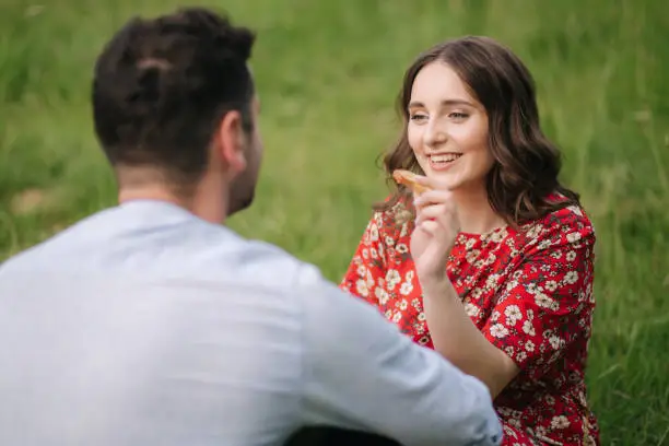 Happy couple spend time outdoors on mini picnic. Attractive woman and handsome man sits on red blanker by the big tree.