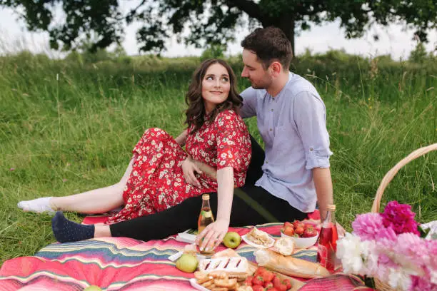 Happy couple spend time outdoors on mini picnic. Attractive woman and handsome man sits on red blanker by the big tree.