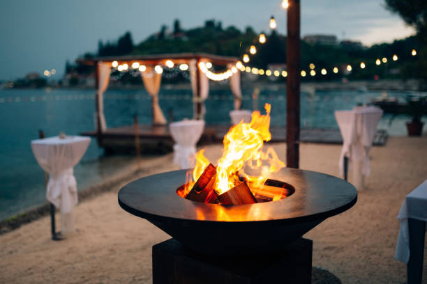 griglia con le fiamme all'interno. superficie di cottura rotonda. sulla spiaggia, sullo sfondo del gazebo vicino all'acqua con ghirlande, nella luce del crepuscolo. - beach sunset sand wood foto e immagini stock