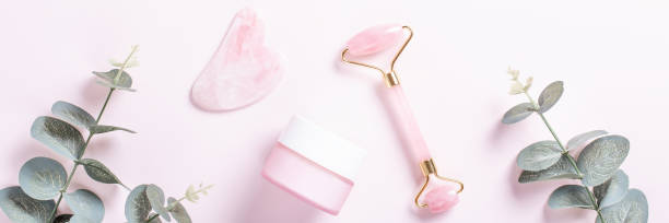 istock Facial roller, gua sha and moisturizing cream with green leaves, web banner. Facial tools on pink. Rose quartz beauty roller, gua sha flat lay. Tools for skin care routines, top view 1253076127