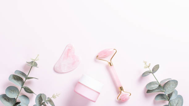 istock Facial roller, gua sha and moisturizing cream with green leaves, web banner. Facial tools on pink. Rose quartz beauty roller, gua sha flat lay. Tools for skin care routines, top view 1253076135