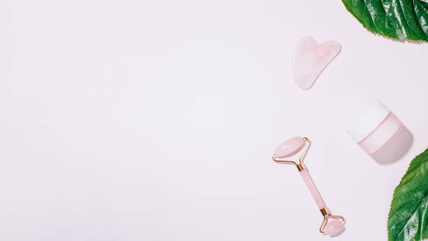 istock Facial roller, gua sha and moisturizing cream with green leaves, web banner. Facial tools on pink. Rose quartz beauty roller, gua sha flat lay. Tools for skin care routines, top view 1253076169