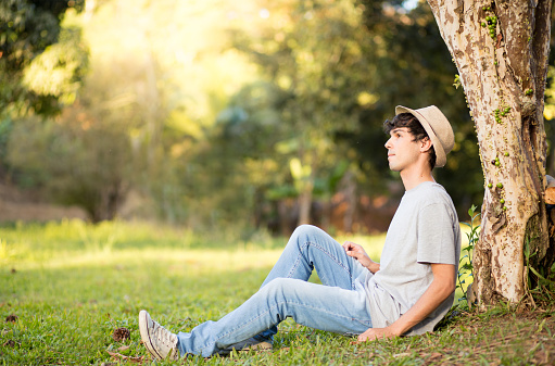 Young man wearing hat sitting under a tree in a public park to relax
