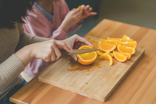 Image of Young Asian mother use kitchen knife cut an orange to pieces with her daughter in living room.