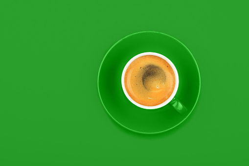 One full espresso coffee cup with saucer over vivid green paper background, elevated top view, directly above