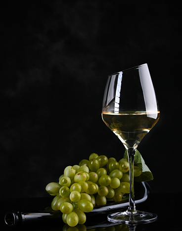 Wine background. White wine in wineglass, grape on old bottle . On the black background.