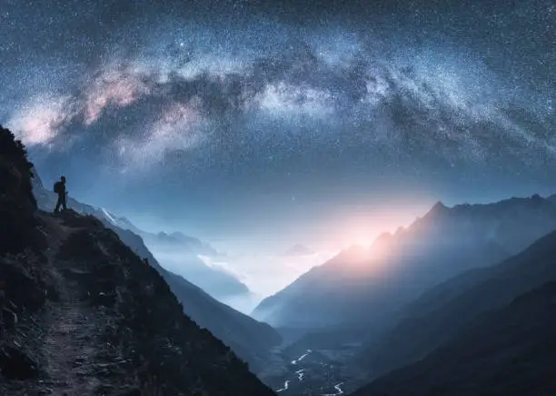 Photo of Arched Milky Way, woman and mountains at night. Silhouette of standing girl on the mountain peak, mountains in low clouds and starry sky in Nepal. Space landscape with bright milky way arch. Travel