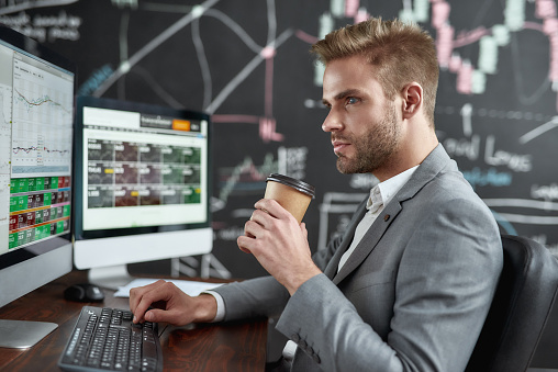 Portrait of successful young trader looking aside and drinking coffee while sitting in front of multiple monitors in the office. Focus on a man. Stock trading, people, business concept. Side view