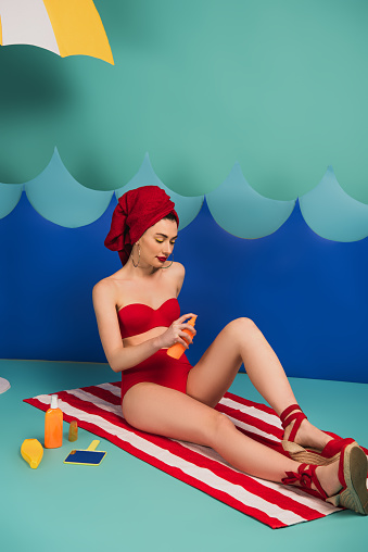 attractive woman in red towel and swimsuit applying sunscreen near paper cut waves