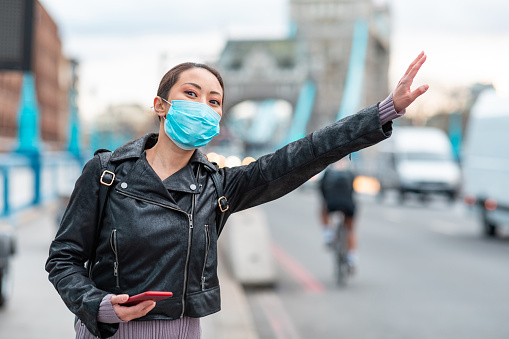 Mujer china en Londres con máscara facial y parar un taxi - joven mujer asiática al lado de la carretera ocupada en la ciudad utilizando un teléfono inteligente para reservar un paseo - conceptos de salud y estilo de vida photo
