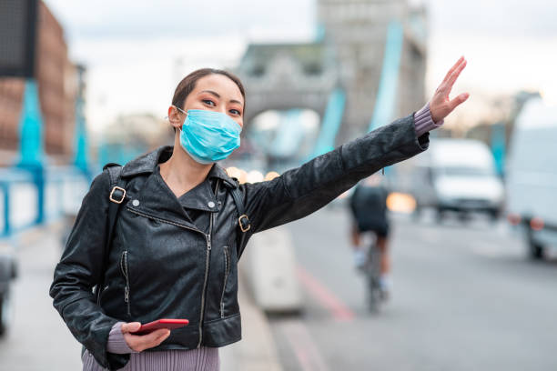 chinese woman in london wearing face mask and hailing a taxi cab - young asian woman next to busy road in the city using a smartphone to book a ride - health and lifestyle concepts - fazendo sinal com a mão imagens e fotografias de stock