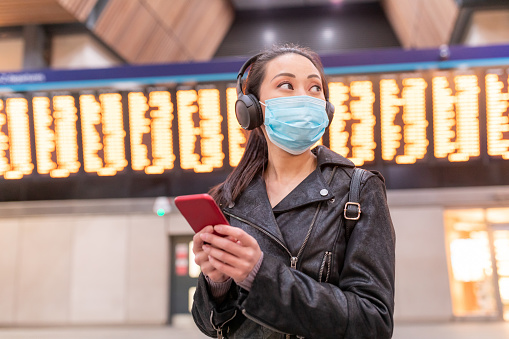 Mujer china que lleva máscara facial en la estación de tren y mantener la distancia social - joven mujer asiática usando el teléfono inteligente y mirando hacia otro lado con la tabla de llegadas de salida detrás - conceptos de salud y viajes photo
