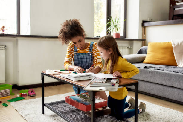 dedication. caucasian little girl spending time with african american baby sitter. they are drawing, learning to write letters, sitting on the floor - nanny imagens e fotografias de stock