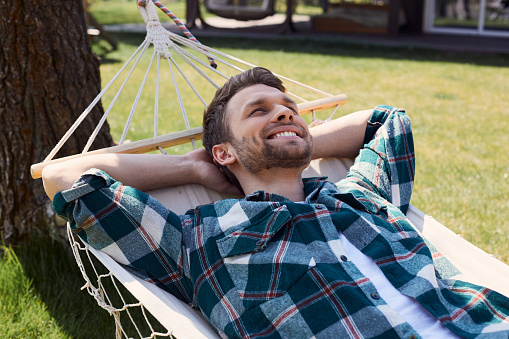 Joyful bearded male looking upwards and keeping hands behind his head while resting on swinging bed