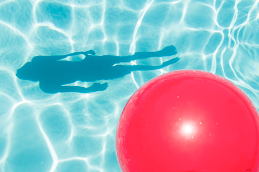 This bright red ball is sitting on the surface of clear blue water in a swimming pool.  Sunlight bouncing from the water's surface is seen reflecting on the bottom of the pool.  The shadow of a passing boy swimmer is also reflected from the bottom of the pool.