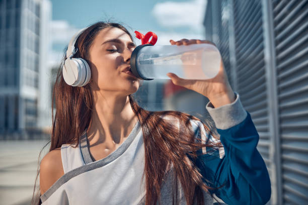 Sportswoman in headphones holding a plastic bottle Close up portrait of a young long-haired stylish female drinking water with her eyes closed outdoors drinking stock pictures, royalty-free photos & images