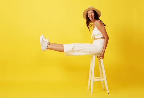Beautiful, crazy young woman with straw hat sitting on white chair front of yellow background. looking at camera. smiling.