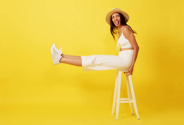 Beautiful, crazy young woman with straw hat sitting on white chair front of yellow background. Beautiful, crazy young woman with straw hat sitting on white chair front of yellow background. looking at camera. smiling. full body isolated stock pictures, royalty-free photos & images